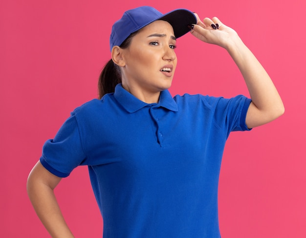Young delivery woman in blue uniform and cap looking aside puzzled standing over pink wall