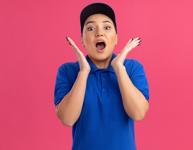 Young delivery woman in blue uniform and cap looking at front amazed and surprised with arms near face standing over pink wall