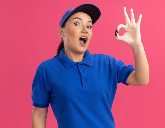 Young delivery woman in blue uniform and cap looking at front happy and excited showing ok sign standing over pink wall