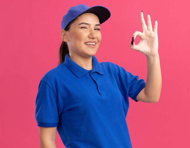 Young delivery woman in blue uniform and cap looking at front smiling cheerfully showing ok sign standing over pink wall