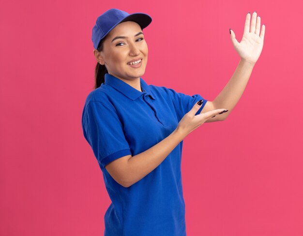 Молодая женщина-доставщик в синей униформе и кепке, глядя вперед, уверенно улыбается, представляя пространство для копирования с руками, стоящими над розовой стеной