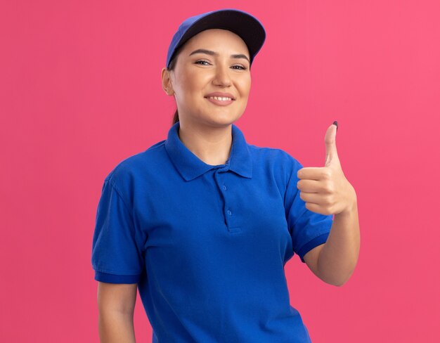 Молодая женщина-доставщик в синей униформе и кепке, глядя вперед, улыбается, уверенно показывает палец вверх, стоя над розовой стеной