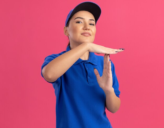 Молодая женщина-доставщик в синей форме и кепке смотрит вперед с уверенным выражением лица, делая жест тайм-аута руками, стоящими над розовой стеной