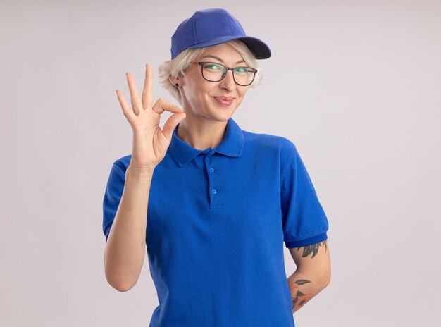 Молодая женщина-доставщик в синей униформе и кепке, выглядящая улыбающейся, уверенно швоинг ок, знак, стоящий над белой стеной