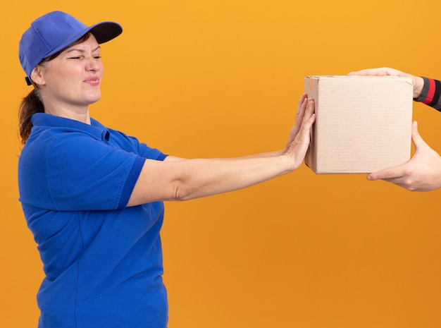 Giovane donna delle consegne in uniforme blu e berretto che sembra preoccupato con un'espressione disgustata che rifiuta di prendere il pacchetto in piedi sopra la parete arancione