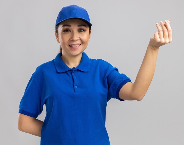 Молодая женщина-доставщик в синей форме и кепке делает денежный жест, потирая пальцы, весело улыбаясь, стоя над белой стеной