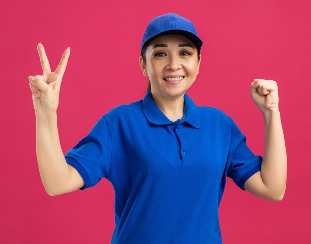 Молодая женщина-доставщик в синей форме и кепке весело улыбается, показывая знак v, сжимающий кулак, стоящий над розовой стеной