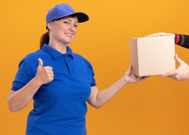 Молодая женщина-доставщик в синей форме и кепке дружелюбно улыбается, показывая знак ОК, получая пакет коробки, стоящий над оранжевой стеной