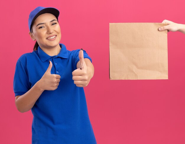 Young delivery woman in blue uniform and cap smiling friendly showing thumbs up while receiving paper package standing over pink wall