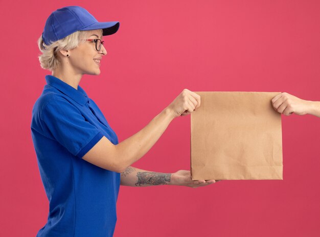 Молодая женщина-доставщик в синей форме и кепке дружелюбно улыбается, получая бумажный пакет, стоящий над розовой стеной