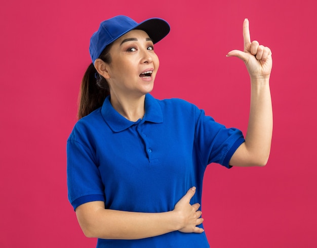Молодая женщина-доставщик в синей форме и кепке улыбается, указывая указательным пальцем вверх, имея новую идею