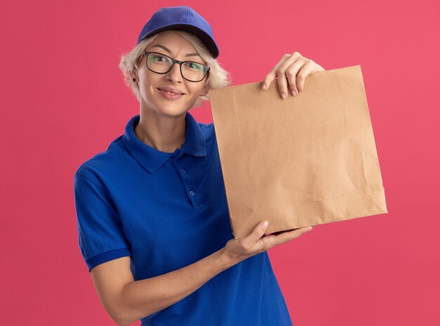 ピンクの壁に元気に笑っている紙のパッケージを保持している眼鏡と青い制服を着た若い配達の女性