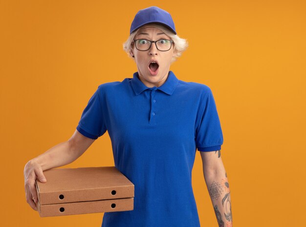 Young delivery woman in blue uniform and cap wearing glasses holding pizza boxes  amazed and surprised over orange wall