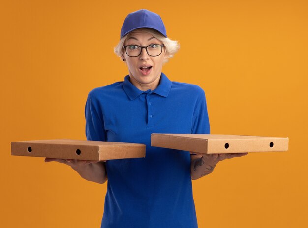 Young delivery woman in blue uniform and cap wearing glasses holding pizza boxes  amazed and surprised over orange wall