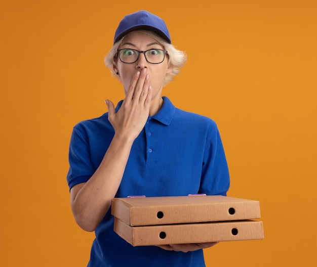 Giovane donna di consegna in uniforme blu e berretto con gli occhiali che tengono le scatole della pizza che è scioccata che copre la bocca con la mano sulla parete arancione