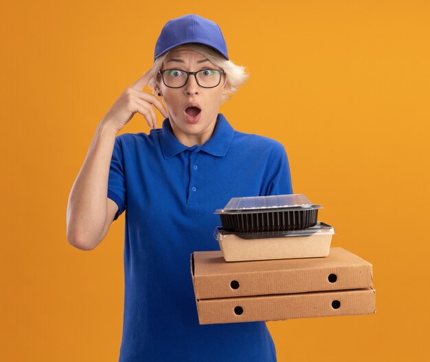 Young delivery woman in blue uniform and cap wearing glasses holding pizza boxes and food packages  surprised over orange wall