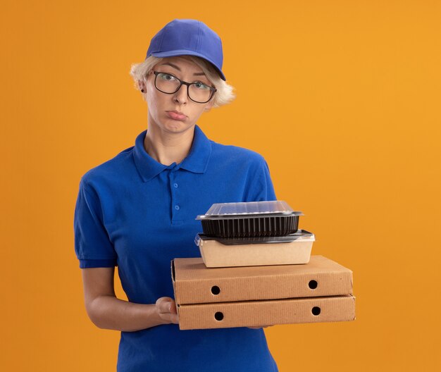青い制服を着た若い配達の女性とオレンジ色の壁の上の顔に悲しい表情でピザボックスと食品パッケージを保持している眼鏡をかけているキャップ