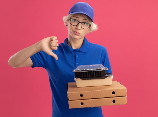 Young delivery woman in blue uniform and cap wearing glasses holding pizza boxes and food packages  with sad expression showing thumbs down over pink wall