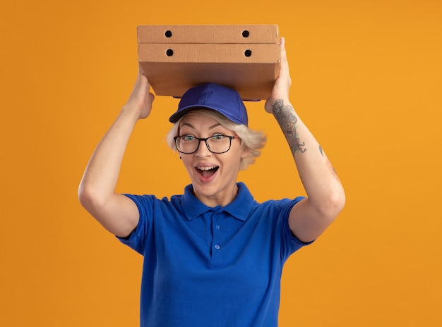青い制服を着た若い配達の女性とオレンジ色の壁に幸せそうな顔で笑って彼女の頭の上にピザの箱を保持している眼鏡をかけているキャップ