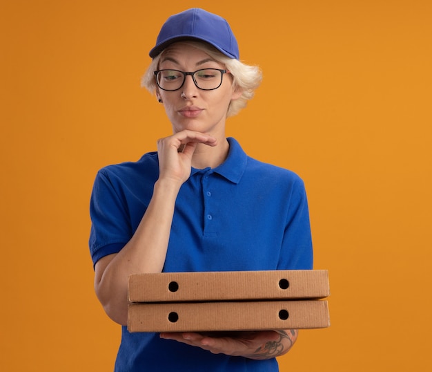 Молодая женщина-доставщик в синей форме и кепке в очках держит коробки для пиццы, задумчиво глядя в сторону над оранжевой стеной