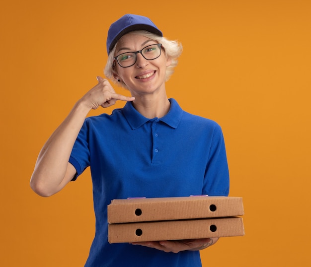 Giovane donna di consegna in uniforme blu e berretto con gli occhiali che tengono le scatole per pizza sorridendo facendomi calare il gesto sorridente sopra la parete arancione