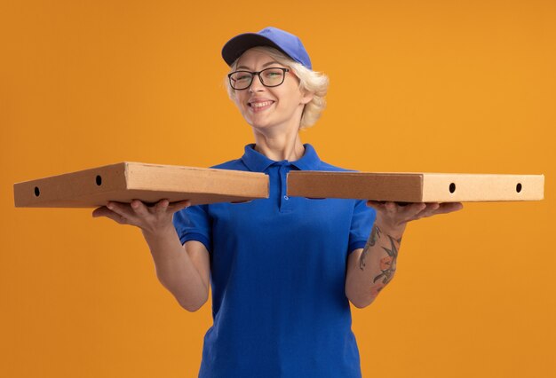 Молодая женщина-доставщик в синей форме и кепке в очках держит коробки для пиццы с большой улыбкой на лице над оранжевой стеной