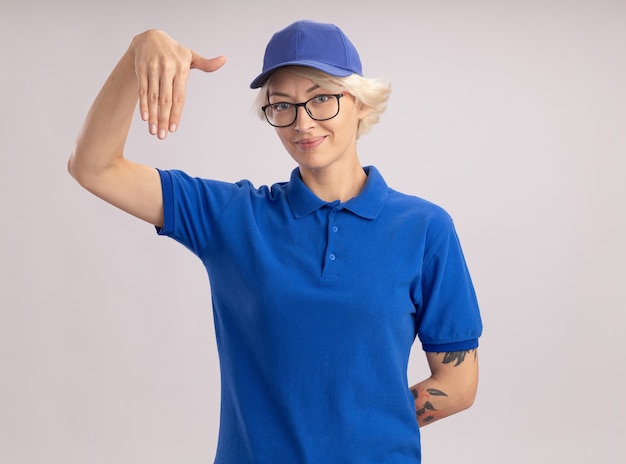 Молодая женщина-доставщик в синей форме и кепке в очках, уверенно выглядящая, жестикулирующая руками, концепция языка тела, стоящая над белой стеной