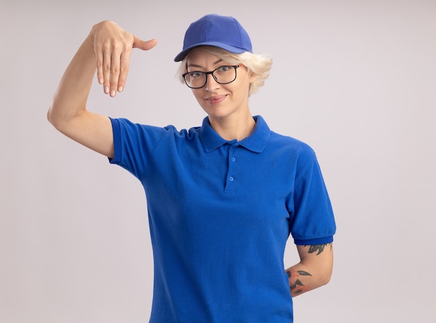 Young delivery woman in blue uniform and cap wearing glasses looking confident gesturing with hands, body language concept standing over white wall