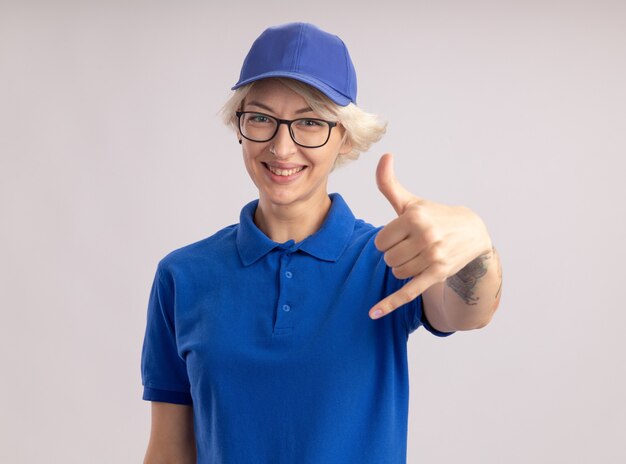 파란색 제복을 입은 젊은 배달 여자와 모자를 쓰고 행복 한 얼굴로 웃고있는 모자를 쓰고 흰 벽 위에 서있는 제스처를 불러주세요.