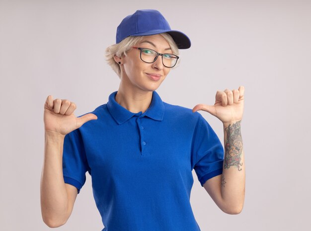 Молодая женщина-доставщик в синей форме и кепке в очках, указывая большими пальцами на себя, стоящую над белой стеной