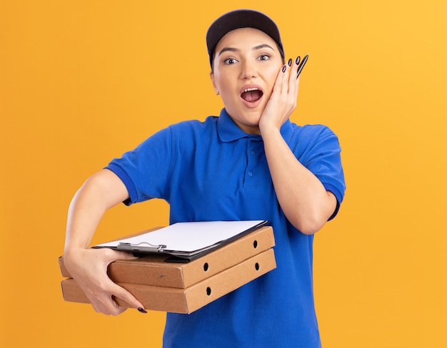 Young delivery woman in blue uniform and cap with clipboard holding pizza boxes looking at front happy and surprised standing over orange wall