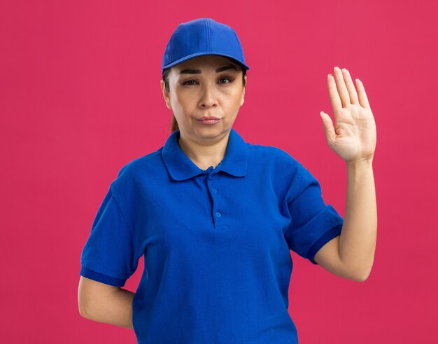 Young delivery woman in blue uniform and cap  with serious face raising hand standing over pink wall