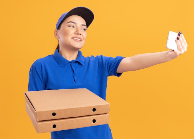 무료 사진 파란색 제복을 입은 젊은 배달 여자와 오렌지 벽 위에 유쾌하게 서있는 셀카를하고 스마트 폰을 사용하여 피자 상자를 들고 모자