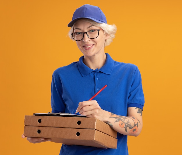 Бесплатное фото Молодая женщина-доставщик в синей форме и кепке в очках держит коробки для пиццы и буфер обмена с пустыми страницами, что-то пишет карандашом, улыбаясь над оранжевой стеной