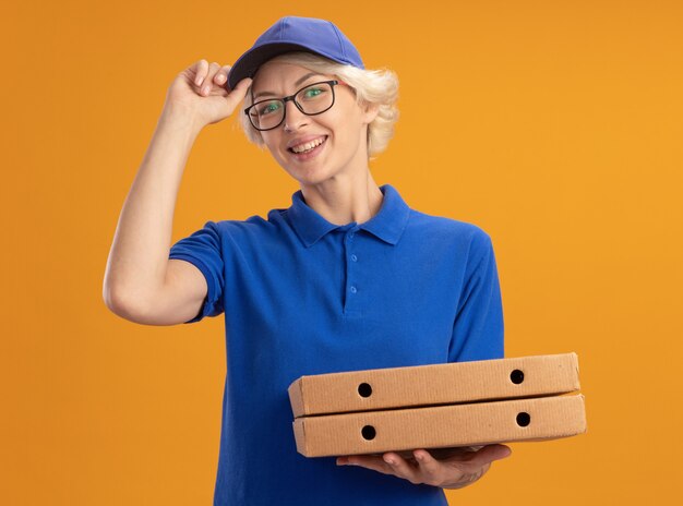 青い制服を着た若い配達の女性とオレンジ色の壁に彼女のキャップを固定する自信を持って笑顔のピザボックスを保持している眼鏡をかけているキャップ