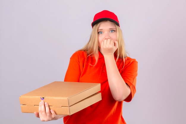 Young delivery woman in orange polo shirt and red cap holding a stack of pizza boxes looking stressed and nervous with hands on mouth biting nails over isolated white background