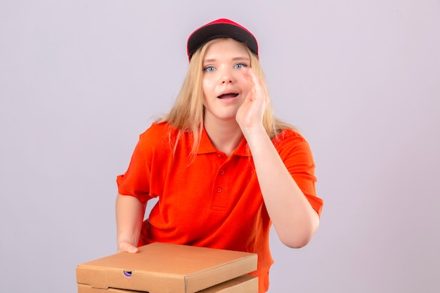 Young delivery woman in orange polo shirt and red cap standing with pizza boxes holding hand near open mouth and saying something over isolated white background