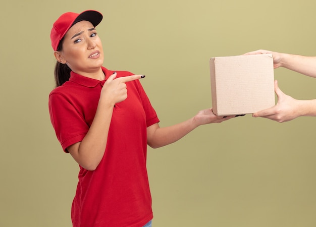 Giovane donna delle consegne in uniforme rossa e cappuccio che si sente confusa mentre riceve il pacchetto della scatola che punta con il dito indice alla scatola che sta sopra la parete verde