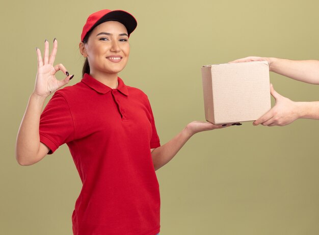 Молодая женщина-доставщик в красной форме и кепке дает картонную коробку клиенту, дружелюбно улыбаясь, показывая знак ОК, стоящий над зеленой стеной