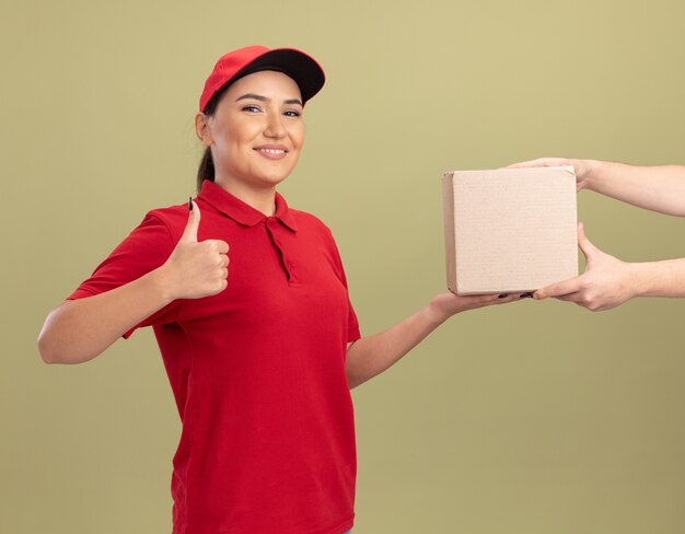 Молодая женщина-доставщик в красной форме и кепке дает картонную коробку клиенту, дружелюбно улыбаясь, показывая пальцы вверх, стоя над зеленой стеной