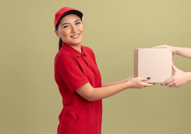 Young delivery woman in red uniform and cap giving cardboard box to a customer smiling friendly standing over green wall