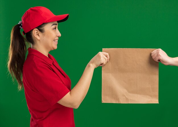 Young delivery woman in red uniform and cap giving paper package to a customer smiling confident standing over green wall