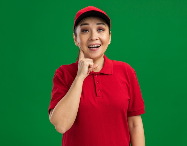 Young delivery woman in red uniform and cap  happy and cheerful smiling standing over green wall
