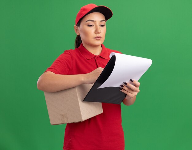 Young delivery woman in red uniform and cap holding cardboard box and clipboard with blank pages looking at it with serious face standing over green wall