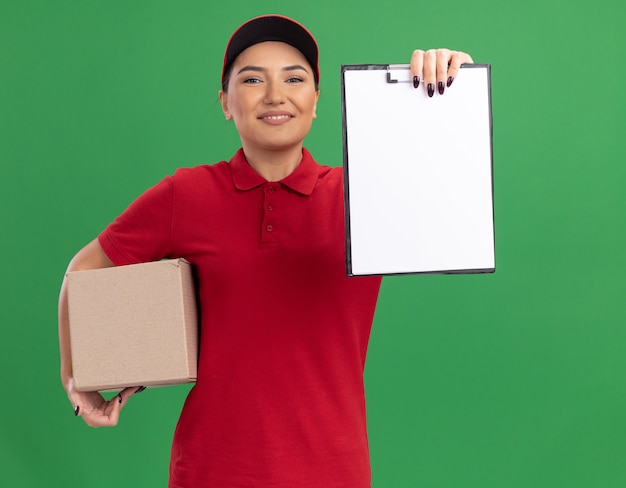 Young delivery woman in red uniform and cap holding cardboard box showing clipboard with blank pages looking at front smiling cheerfully standing over green wall