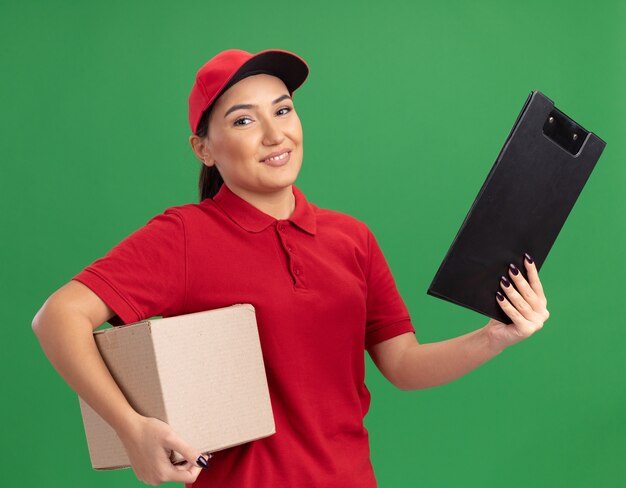 녹색 벽 위에 자신감 서 웃 고 행복 한 얼굴로 앞을보고 클립 보드와 골 판지 상자를 들고 빨간 유니폼과 모자 젊은 배달 여자