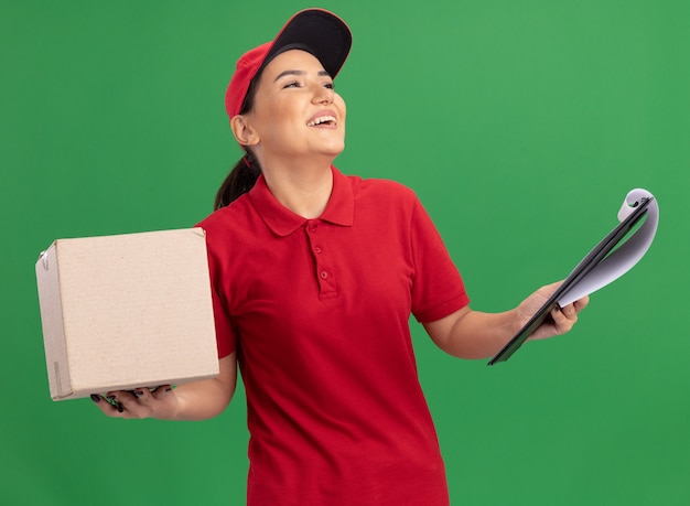 Giovane donna di consegna in uniforme rossa e cappuccio che tiene la scatola di cartone con appunti che osserva in su con il sorriso sul fronte che sta sopra la parete verde