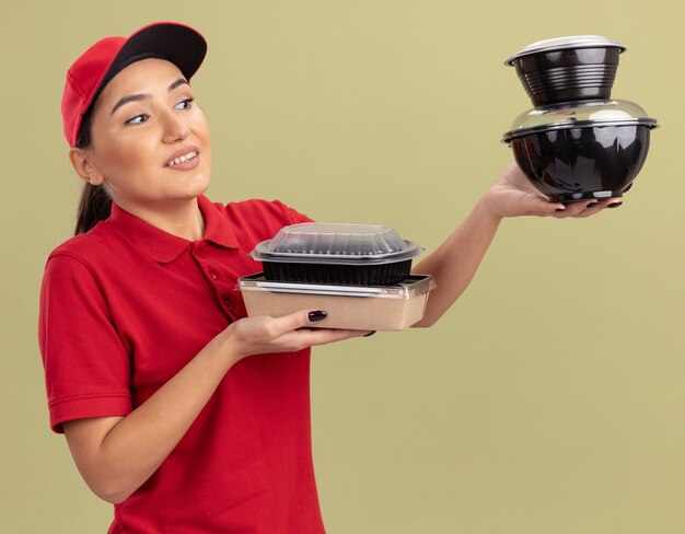 young delivery woman in red uniform and cap holding food packages looking at them smiling with happy face standing over green wall