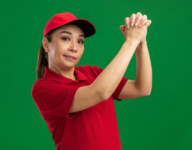 緑の壁の上に立つ自信のある表情でチームワークのジェスチャーをする赤い制服と帽子を着た若い配達女性
