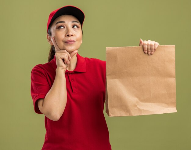 赤い制服を着た若い配達女性と、緑の壁の上に立って当惑して見上げる紙のパッケージを持ったキャップ